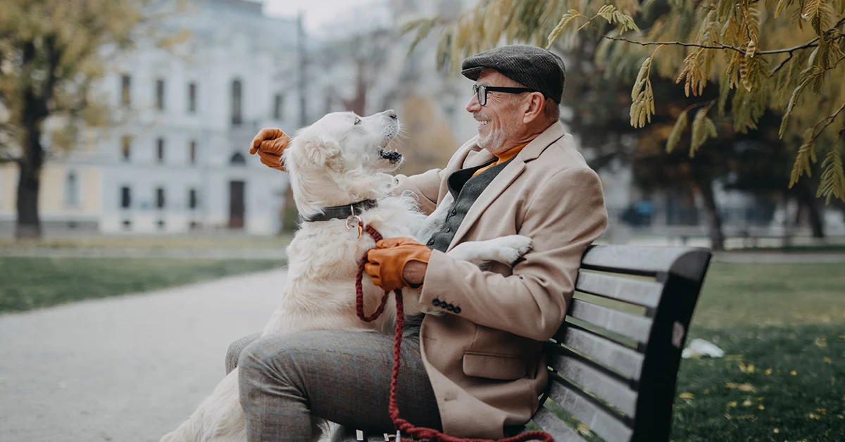 A smiling older man wearing a flat cap and tan overcoat sits on a park bench, happily hugging a large, white dog that is looking up at him.