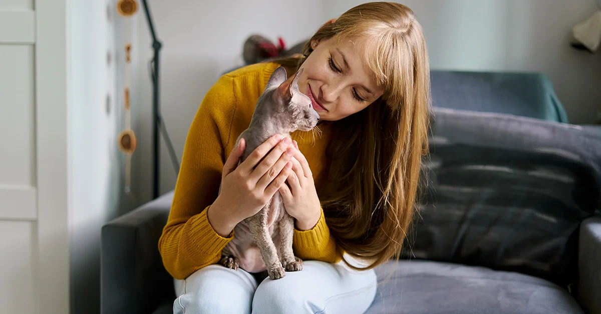 A smiling woman in a yellow sweater sits on a sofa, gently cradling a Sphynx cat in her lap.