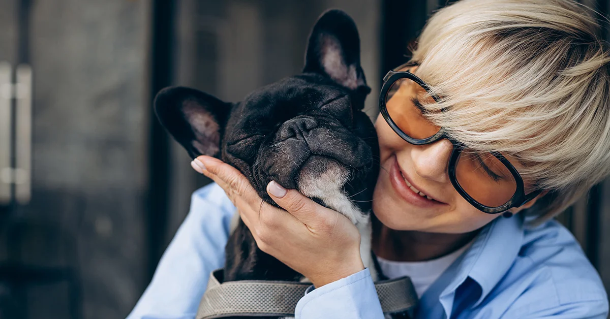 happy-frenchie-cuddles A smiling person with short blonde hair affectionately cradles a black French Bulldog's head.