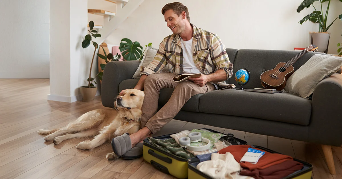 A man in a plaid shirt sits on a dark grey sofa, smiling as he pets his Golden Retriever.