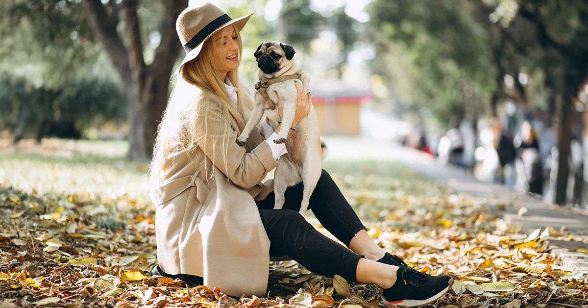 A woman in a hat sitting in a park and holding a pug.