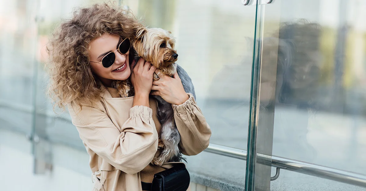 A smiling woman with curly hair and sunglasses holding a small long-haired dog.